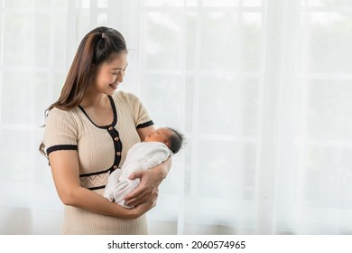 Beautiful Asian Woman Holding Newborn Baby In Her Arms Standing In Front Of Windows At Cozy Home.Happy Infant Baby Sleep In Mother Arms Safety And Comfortable.Mom And Baby Concept