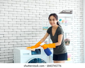 Beautiful Asian Woman, Housewife Wearing Orange Protective Rubber Gloves Using Cleaner Spray And Clean Cloth To Cleaning Washing Machine On White Brick Wall Background At Laundry Room With Copy Space.