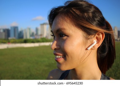 Beautiful Asian Woman Listening To Her Air Pods In Sports Attire On A Grass Field Near The City