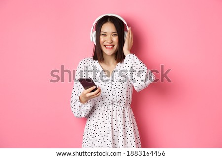 Beautiful asian woman listening music in headphones, using smartphone streaming app and smiling, standing over pink background