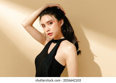 Beautiful Asian woman with long curly hair and green eyes in a black sexy swimsuit on a isolated cream background. Young female model with perfect skin posing in fashion while looking at the camera.