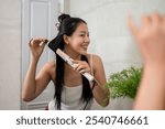 A beautiful Asian woman with long, natural black hair is curling her hair with a curling iron in front of the bathroom mirror, smiling at her reflection.