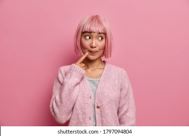 Beautiful Asian Woman Looks With Surprised Curious Expression On Right Side, Keeps Index Finger Near Mouth, Has Dyed Rosy Hair, Models Indoor, Dressed In Comfortable Sweater. Thoughtful Charming Lady