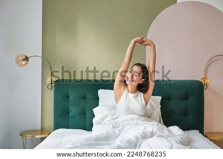 Beautiful asian woman lying in her bedroom on bed, stretching hands and looking outside, waking up from sleep.