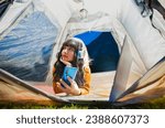 Beautiful asian woman lying inside tent with wireless headphones on her head, use mobile phone and laptop while camping in the beautiful countryside at night and looking through the tent. 