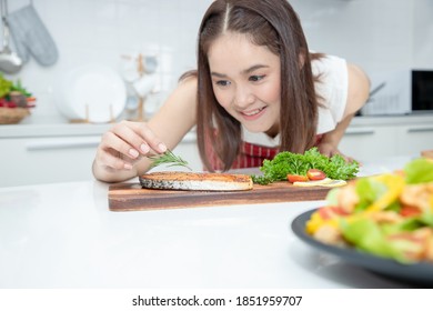 A beautiful Asian woman places rosemary on a salmon steak arranged on a plate with green oak vegetables. Ideas about healthy cooking and weight loss. Soft focus - Shutterstock ID 1851959707