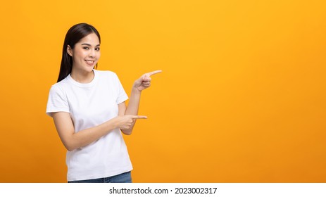 Beautiful asian woman pointing finger into blank space. Smiling female wearing white shirt on isolated yellow background. Attractive Teenager girl looking at empty space for text.