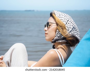 Beautiful Asian Woman Portrait With Closing Eyes In Casual White Shirt Wearing Sunglasses And Hair Scarf Sit On Blue Bean Bag Seat On The Beach With Sea View And Blue Sky Background In Summer.