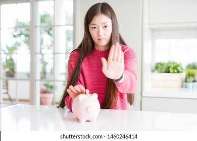 Beautiful Asian woman putting a coin inside piggy bag with open hand doing stop sign with serious and confident expression, defense gesture Stock Photo