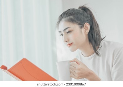 beautiful asian woman resting in the room On weekends, she has big eyes. and floating a beautiful smile. she holds a white glass And there was an orange book in her hand. - Powered by Shutterstock