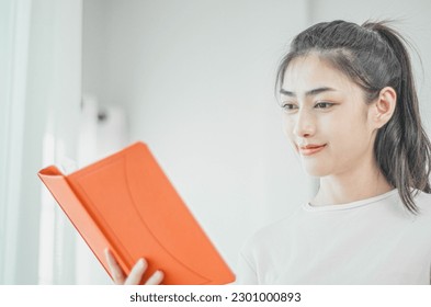 beautiful asian woman resting in the room On weekends, she has big eyes. and floating a beautiful smile. she holds a white glass And there was an orange book in her hand. - Powered by Shutterstock