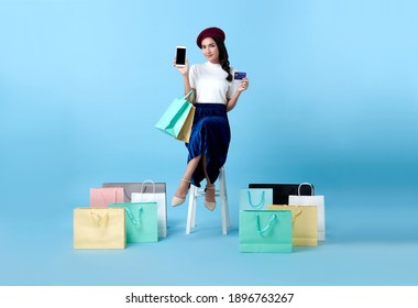 Beautiful Asian Woman Shopper Sitting And Carrying Shopping Bags With Showing Mobile Phone And Credit Card In Hands On Blue Background.