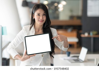 Beautiful Asian Woman Showing Blank Screen Tablet While Standing In Modern Office Room, Smiling, Looking At Camera And Pointing At Tablet Screen.