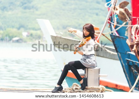 Beautiful asian woman sit,hold and play violin on sea view background near big ship, in emotion feeling happy.