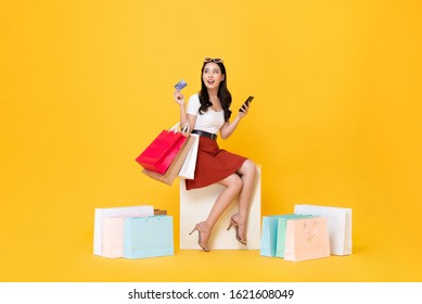 Beautiful Asian Woman Sitting And Carrying Shopping Bags With Credit Card And Mobile Phone In Hands On Yellow Background