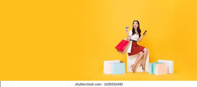 Beautiful Asian Woman Sitting And Carrying Shopping Bags With Credit Card And Mobile Phone In Hands On Yellow Banner Background