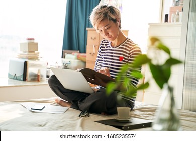 Beautiful Asian Woman Sitting On Bed & Working From Home On Laptop