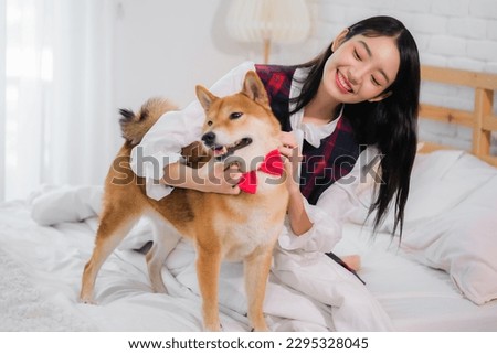 Beautiful asian woman sitting on white bed in bedroom while scratching dog in playful expression.