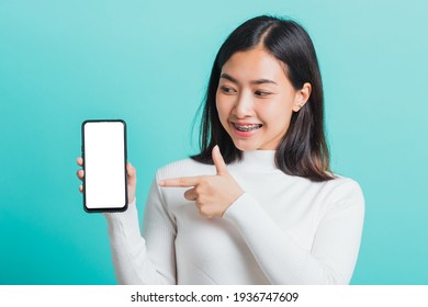 Beautiful Asian Woman Smile Holding A Smartphone On Hand And Pointing Finger To The Blank Screen, Female Excited Cheerful Her Show Mobile Phone Isolated On A Blue Background, Technology Concept