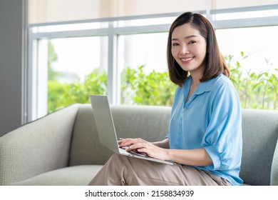 Beautiful Asian Woman Smile Wearing Blue Shirt Using Laptop And Sitting On Sofa In Living Room At Home.Happy Relaxing On Holiday