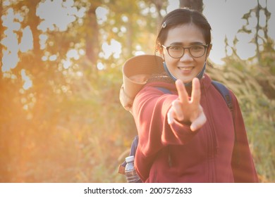 

Beautiful Asian Woman Smiling Happily Looking Straight Up And Holding Up Two Fingers With A Water Bottle Backpack. While Out Hiking On Holiday Tropical Hills And The Orange Light Of The Morning Sun.