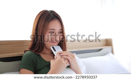Beautiful Asian Woman using mobile phone enjoying chatting, surfing on the internet on a phone on bed. Happy woman looking at smartphone texting messages, chatting and smile at bedroom