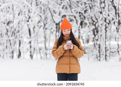 Beautiful Asian Woman Walking In The Park, Uses The Phone For Online Shopping, On A Winter Snowy Day