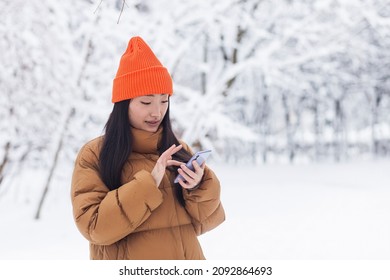 Beautiful Asian Woman Walking In The Park, Uses The Phone For Online Shopping, On A Winter Snowy Day