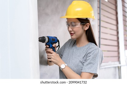 Beautiful Asian Woman Wear Protective Helmet And Glasses Using Electric Drill Improvement During The Installation Of White Wall. Adult Making DIY Workshop. Repairing Home. Moving In New House Concept.