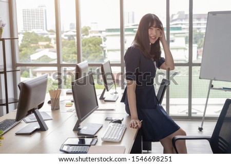 A beautiful Asian woman is wearing a dark blue suit sitting at a desk in a modern office and is happy to work and has a large glass window background.
