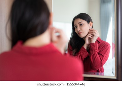 Beautiful Asian Woman Wearing Red Dressed Putting Star Earring Looking In Mirror In Her Bedroom At Home. Makeup In Morning Getting Ready Before Going To Work.