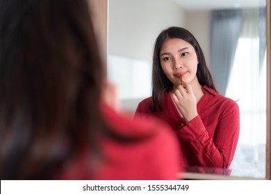 Beautiful Asian Woman Wearing Red Dressed Putting Lipstick Looking In Mirror In Her Bedroom At Home. Makeup In Morning Getting Ready Before Going To Work.