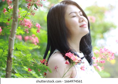 Beautiful Asian Woman In White Dress Close Her Eyes And Smelling In Blooming Flowers With Natural In Garden Sunlight. Thai Or Chinese Girl Enjoy On Holiday 