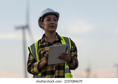 Beautiful Asian woman in white helmet working with digital tablet at renewable energy farm. Female inspector controlling functioning of wind turbines outdoors. - Powered by Shutterstock