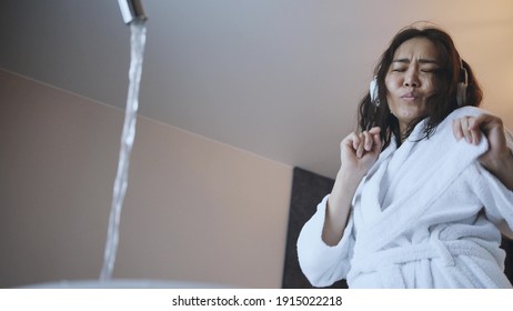 Beautiful Asian Woman In White Robe Listens To Music With Wireless Headphones In The Bathroom.