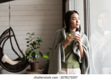 Beautiful asian woman wrapped in blanket, leaning on window and looking outside, drinking hot tea - Powered by Shutterstock