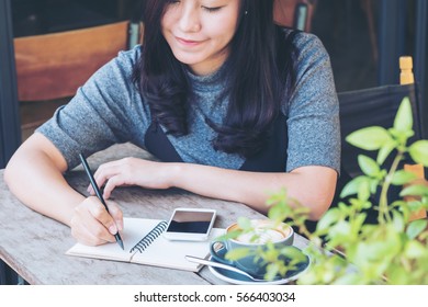 A Beautiful Asian Woman Writing On Notebook With Hot Coffee And Smartphone In Vintage Cafe Background