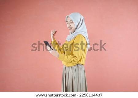Beautiful Asian woman in yellow shirt and hijab smiling cheerful using mobile phone gesturing yes with raised fist on brown background