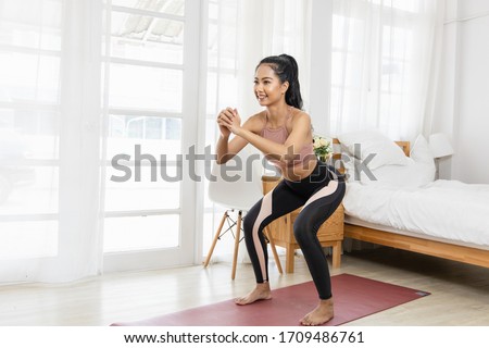 A beautiful Asian woman's fitness at home instead of going to the gym. She is doing squats on a yoga mat in the bedroom. She wears sportswear. Exercise concept for good shape