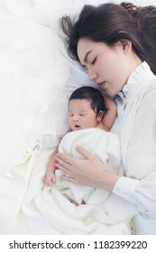 Beautiful Asian Women Mother With Long Black Hair In The White Pajamas. Mom Looking At Newborn Infant With Love, While A Baby Sleeping In Her Arm With Warm, Safe, Comforted Resting On The Clean Bed.