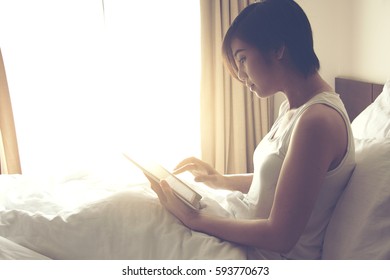 Beautiful Asian women wear white shirt sit on bed and play tablet in the morning at bedroom. - Shutterstock ID 593770673