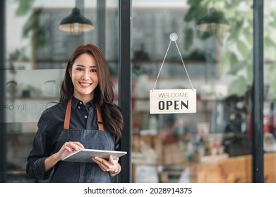 Beautiful asian young barista woman in apron holding tablet and standing in front of the door of cafe with open sign board. Business owner startup concept. - Powered by Shutterstock