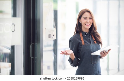 Beautiful Asian Young Barista Woman In Apron Holding Tablet And Standing In Front Of The Door Of Cafe With Open Sign Board. Business Owner Startup Concept.