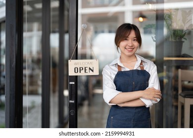 Beautiful Asian Young Barista Woman In Apron Holding Tablet And Standing In Front Of The Door Of Cafe With Open Sign Board. Business Owner Startup Concept