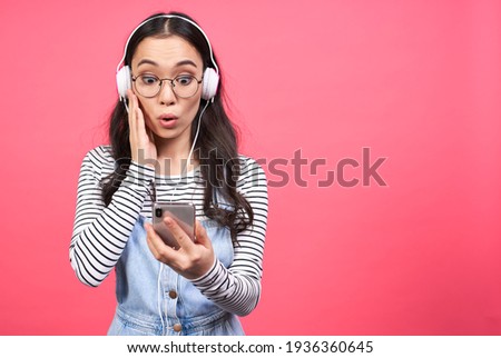 A beautiful Asian young lady receives a funny video from a friend, looks at a mobile phone in surprise, listens to the sound in headphones, is wearing a striped sweater, listens to music. Copy space.