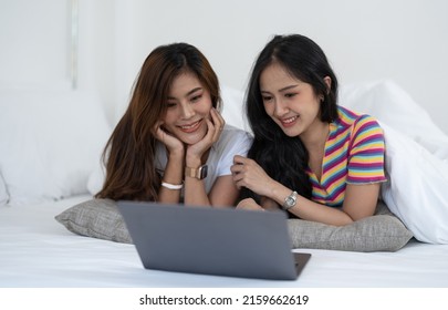 Beautiful Asian Young Lesbian LGBTQ Couple Or Friends Using Computer Laptop Together Smile And Relax On Bed.Happiness LGBTQ Couple Woman Spending Time Together At Home. LGBTQ Pride Month Concept