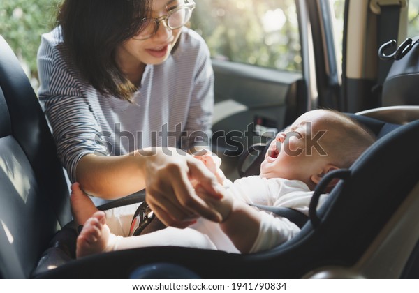 Beautiful Asian\
young mother comforting her baby boy crying while putting and\
fasten seat belts on his car seat in\
car.