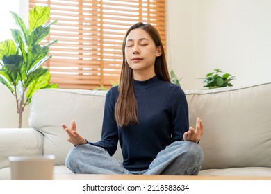 Beautiful Asian Young Woman, Girl Hands In Calm Pose Sitting Practice Meditating In Lotus Position On Sofa At Home, Meditation, Exercise For Wellbeing, Healthy Care. Relaxation, Happy Leisure People.