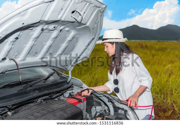 Beautiful Asian young\
woman have problem with battery power of car. she connecting cables\
car battery for recharge the battery car. Automotive motor problem\
concept.