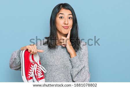 Beautiful asian young woman holding red casual shoes serious face thinking about question with hand on chin, thoughtful about confusing idea 